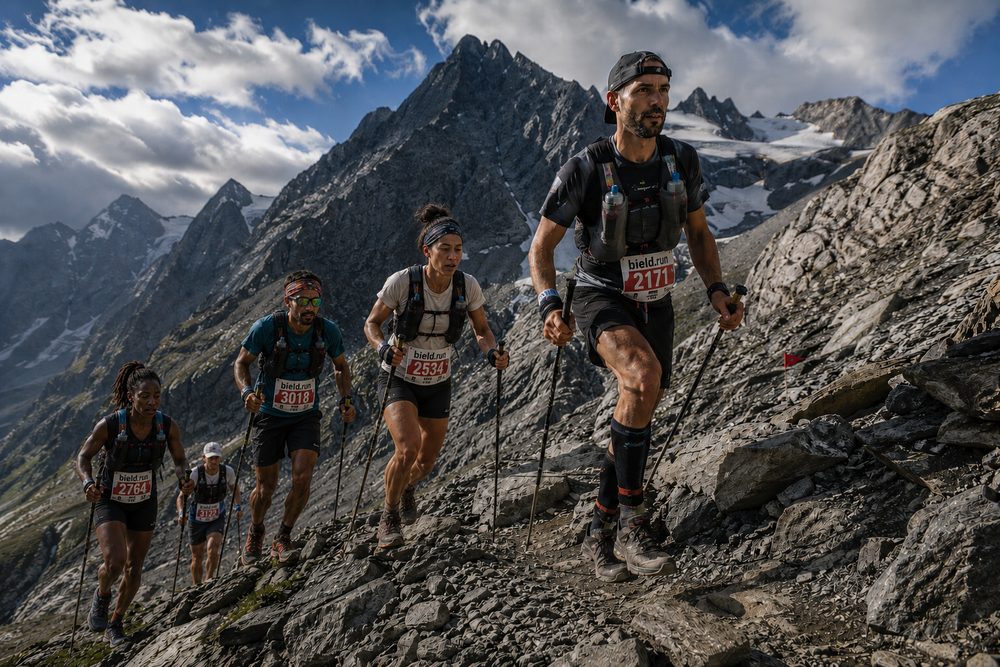 Endurance runners climbing a rocky alpine trail with trekking poles and race bibs, snow-capped peaks behind